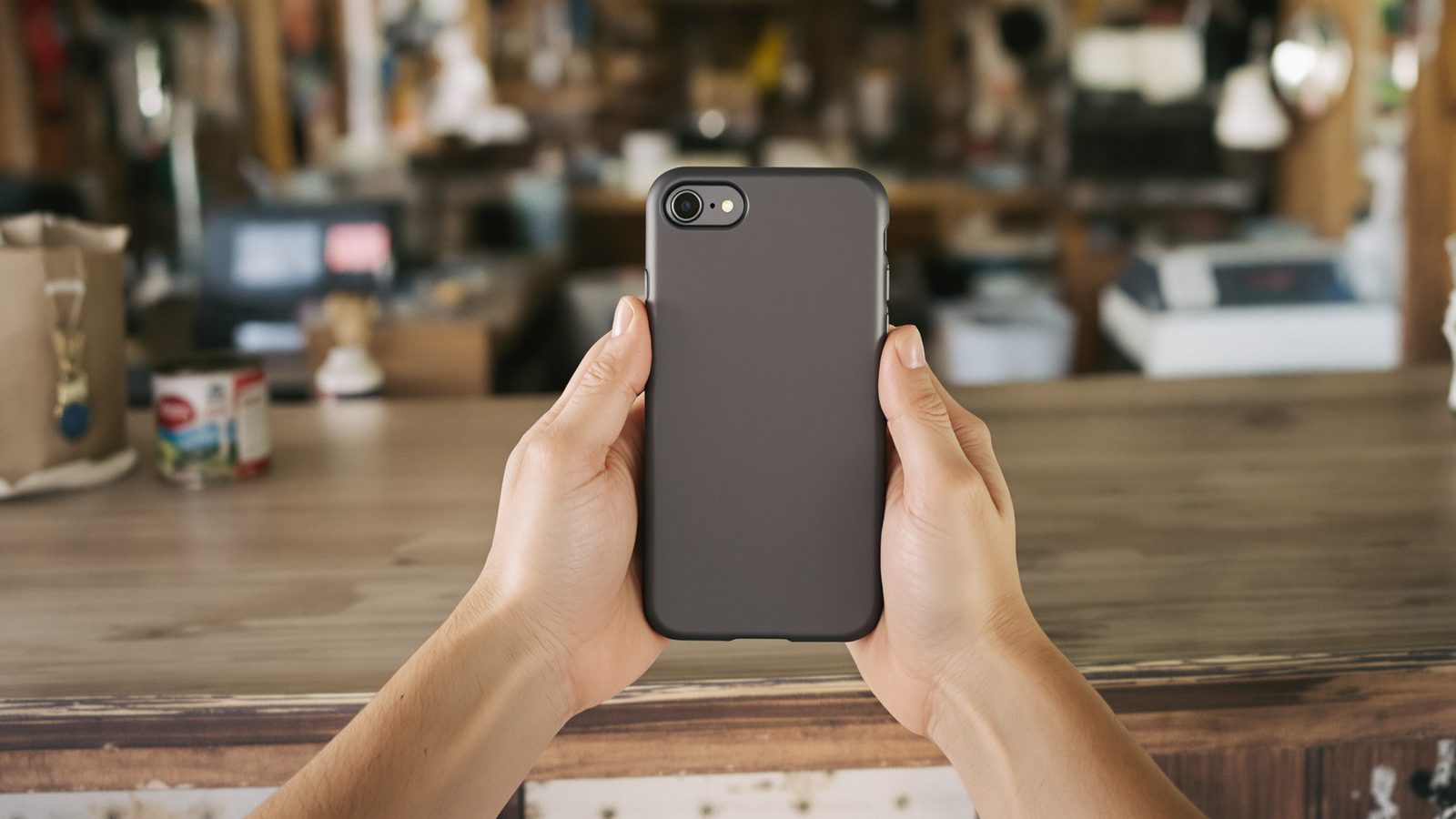 Two hands holding a smartphone above a wooden store counter in soft window light.